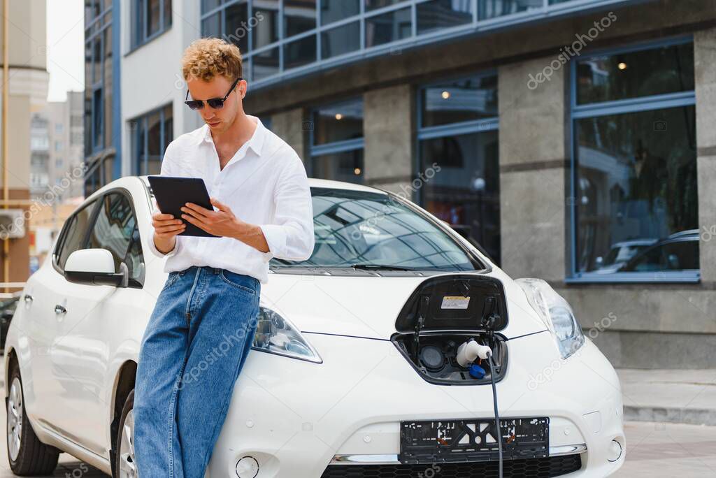 portrait of young handsome man in casual wear, standing at the charging station. Eco electric car concept