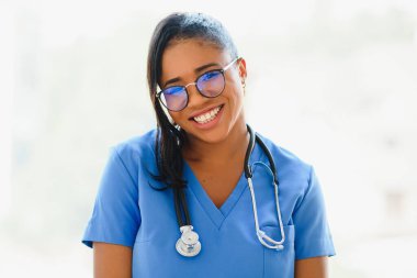 Dost canlısı, kendine güvenen, laboratuvar önlüklü, kollarını çapraz bağlamış gözlüklü bir kadın sağlık uzmanı portresi. Tecrit edilmiş hastane kliniği geçmişi.