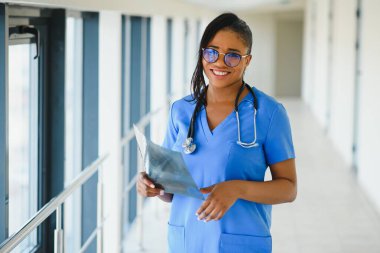 Dost canlısı, kendine güvenen, laboratuvar önlüklü, kollarını çapraz bağlamış gözlüklü bir kadın sağlık uzmanı portresi. Tecrit edilmiş hastane kliniği geçmişi.