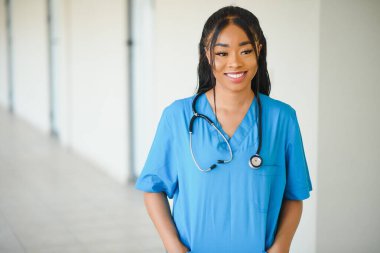 Dost canlısı, kendine güvenen, laboratuvar önlüklü, kollarını çapraz bağlamış gözlüklü bir kadın sağlık uzmanı portresi. Tecrit edilmiş hastane kliniği geçmişi.