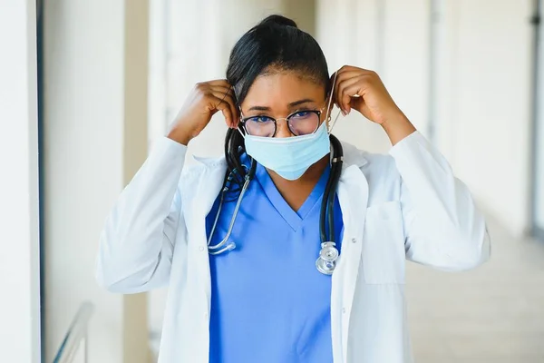 Portrait of african american female doctor wearing mask standing in hospital corridor. medicine, health and healthcare services during coronavirus covid 19 pandemic.