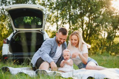 Genç bir aile. Beyaz elbiseli üç kişi piknik yapıyor. Güzel ebeveynler ve kızlar yaz tatilinde arabayla seyahat ederler..