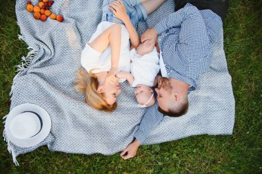 Anne, baba ve küçük oğlu yaz parkında battaniyenin üzerinde yatıyor. Yaz tatili kavramı. Anneler günü, babalar günü, bebekler günü. Aile doğada birlikte vakit geçiriyor. Aile görünüşü
