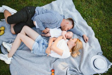 Anne, baba ve küçük oğlu yaz parkında battaniyenin üzerinde yatıyor. Yaz tatili kavramı. Anneler günü, babalar günü, bebekler günü. Aile doğada birlikte vakit geçiriyor. Aile görünüşü