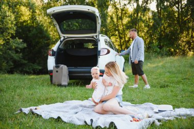 Genç bir aile. Beyaz elbiseli üç kişi piknik yapıyor. Güzel ebeveynler ve kızlar yaz tatilinde arabayla seyahat ederler..