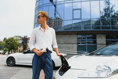 Yakışıklı bir genç adam modern elektrikli arabasını şarj ediyor..