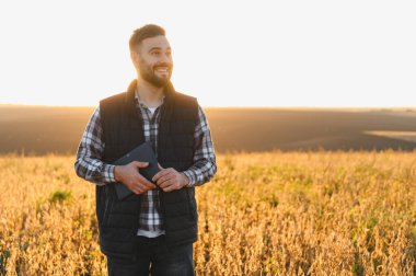 Kırsal bir alanda elinde dijital tabletle dikilen ve ekinleri gözlemleyen genç adam akıllı çiftçiliği temsil ediyor.