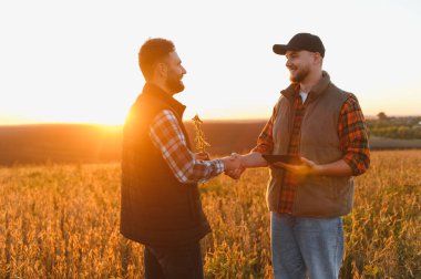 Çiftçiler gün batımında altın bir tarlada el sıkışıyorlar, biri bir ödül tutuyor, diğeri ise dijital tablet.