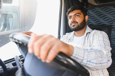 Sakallı adam kamyon kullanıyor, kendinden emin bir şekilde ileriye bakıyor. Uzun bir nakliye aracını temsil ediyor.