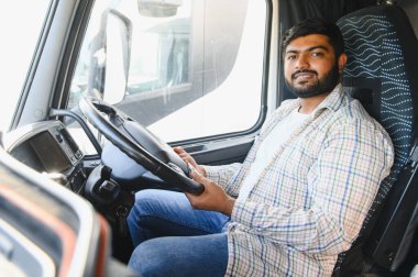 Kareli gömlekli Hintli kamyon şoförü kulübede oturuyor, elleri direksiyonda, işe giderken kameraya güvenle bakıyor.