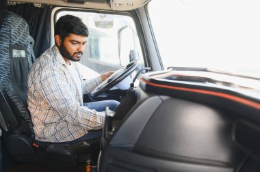 Profesyonel kamyon şoförü olarak çalışan Hintli bir adam. Kamyon kabininde oturuyor ve direksiyonu tutuyor.