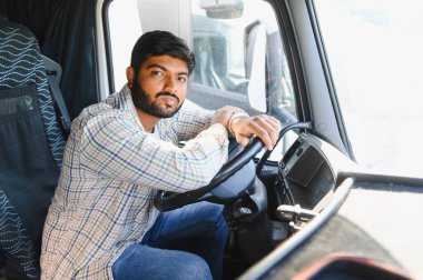 Kamyon kabinindeki genç Hintli adam portresi, direksiyonu güvenle tutuyor, ulaşım ve lojistiği temsil ediyor.