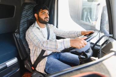 Büyük bir ticari kamyon süren, ekose gömlek ve kot giyen Hintli bir adam, önündeki yola odaklanmış.
