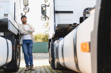 Gülümseyen Hintli adam kollarını kavuşturuyor, beyaz bir tırın yanında duruyor, lojistik ve ulaşımı temsil ediyor.