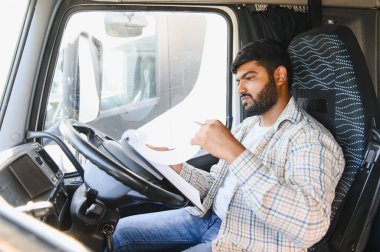Hintli kamyon şoförü kamyon kulübesinde oturuyor, önemli lojistik evraklarını tutuyor ve inceliyor.