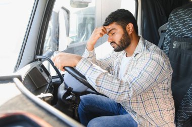 Kamyon şoförü, araç kabininde otururken yorgun ve stresli hissediyor. Uzun yolculukta baş ağrısı çekiyor.