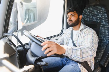 Hintli bir adam büyük bir kargo kamyonu kullanıyor, emniyet kemeri takıyor, yola odaklanıyor, ulaşım ve lojistiği temsil ediyor.