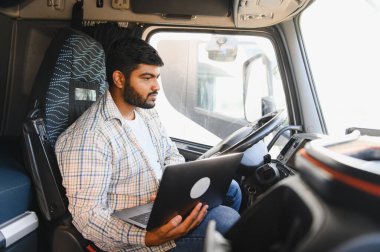 Kamyon kabinindeki Hintli adam dizüstü bilgisayar kullanıyor, iş sırasında lojistiği yönetiyor veya iletişim kuruyor, modern ulaşımı temsil ediyor.