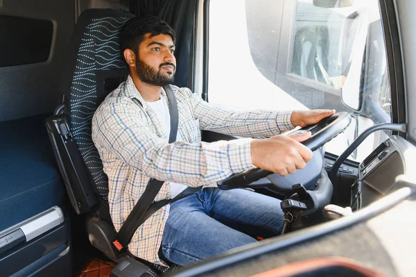 Büyük bir ticari kamyon süren, ekose gömlek ve kot giyen Hintli bir adam, önündeki yola odaklanmış.