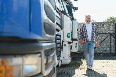 Genç erkek sürücü gülümsüyor, park halindeki ağır yük araçlarında yürüyor, taşımacılık ve lojistik sektörünü temsil ediyor.