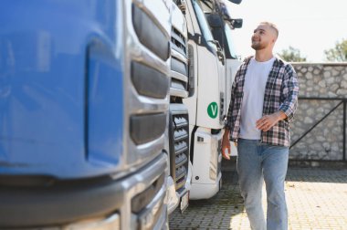 Genç adam, lojistik ve nakliye araçlarını temsil eden ağır görev kamyonlarının yanından güvenle geçiyor ve gülümsüyor.