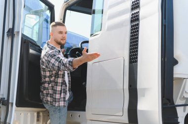 Erkek kamyon şoförü beyaz bir tırın yanında duruyor, eliyle işaret ediyor. Lojistik ve ulaşım temsilcisi