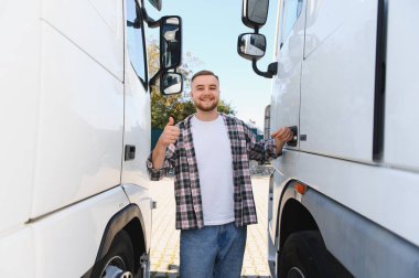 Genç bir adam, profesyonel bir kamyon şoförü, gülümsüyor ve iki büyük nakliye kamyonu arasında bir jest yapıyor.