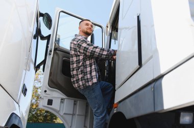 Ekose gömlekli ve kotlu gülümseyen bir adam kamyonun kulübesine giriyor, kendinden emin ve yolda nakliye işi için hazır.