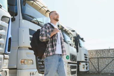 Sırt çantası ve flanel gömlekli genç adam park halindeki kamyonların önünde duruyor, lojistik ve taşımacılığı temsil ediyor.