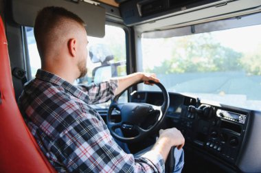 Ekose gömlekli profesyonel bir kamyon şoförü elinde direksiyon, açık bir günde yarı kamyon sürüyor, ilerideki yola odaklanıyor.