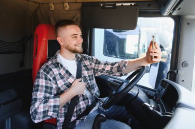 Flanellli gülümseyen kamyon şoförü emniyet kemerini takar ve kabininin içinde kendinden emin ve bağlı bir şekilde park halindeyken akıllı telefon özçekimi çeker.