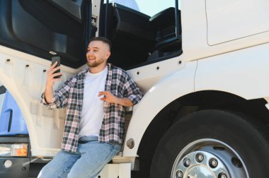 Gülümseyen genç kamyoncu kamyonun kapısında otururken akıllı telefondan bir video görüşmesi yapıyor.