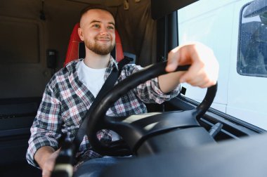 Genç erkek kamyon şoförü takside oturuyor, ekose gömlek ve emniyet kemeri takıyor, direksiyonu sıkıca tutuyor.