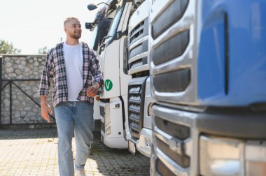 Gülümseyen genç adam, park halindeki ağır kamyonların yanından geçiyor. Taşımacılık, lojistik ve ulaşım endüstrisinin temsilcisi