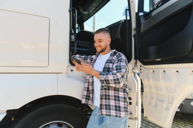 Kabin kapısında dikilen gülümseyen genç adam, ağır bir araç kullanmaya ara verirken telefonu kontrol ediyor.