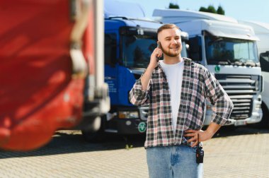 Genç gülen kamyon şoförü telefonla iletişim kuruyor, lojistiği yönetiyor ve tırlarla otoparkta ulaşımı sağlıyor.