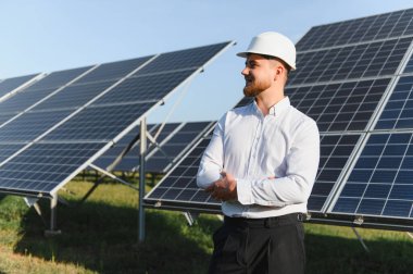 Sert bir şapka takan mühendis, çapraz kollarla, yenilenebilir enerji santralindeki güneş panellerini inceliyor.