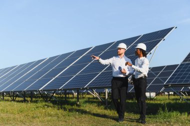 Sert şapka ve üniforma giyen mühendisler güneş panellerini inceliyor, temiz enerji ve sürdürülebilir teknoloji üzerinde işbirliği yapıyorlar.