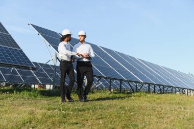 Güneşli bir günde güneş panellerinin önünde durarak yenilenebilir enerji projesini tartışan iki farklı mühendis.