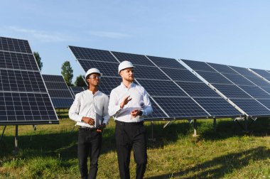 Başlıklı iki mühendis parlak mavi gökyüzü altında bir güneş çiftliğinde yürür ve operasyonları tartışır, panelleri ve altyapıyı incelerler.