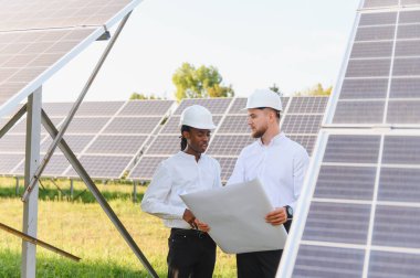 Katı şapkalı mühendisler ellerinde planlar tutuyorlar, güneş panelleri arasında proje planlarını tartışıyorlar, sürdürülebilir enerji gelişimini sembolize ediyorlar