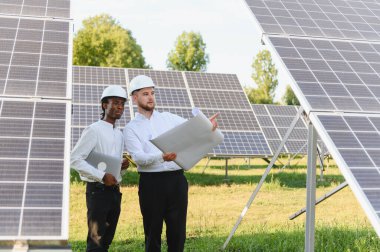 Sert şapkalar takan çeşitli mühendisler güneş paneli açıklarında durup sürdürülebilir enerji planları yaparken planlarını tartışıyorlar.