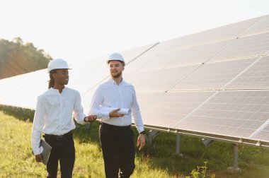 Güneş paneli alanında yürüyen iki farklı erkek mühendis yenilenebilir enerji altyapı projesini tartışıyorlar.