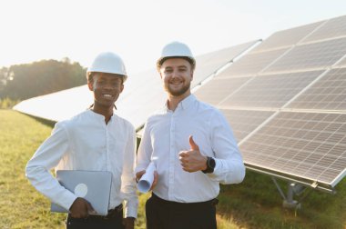Gün batımında güneş paneli alanında duran iki farklı mühendis gülümsüyor, yeşil enerjiyle takım çalışmasını temsil ediyorlar.