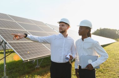 Güvenlik kaskı takan mühendisler, sürdürülebilir yeşil enerjiyi yönetmek için birçok güneş panelinin yanında dururken projeyi tartışıyorlar.