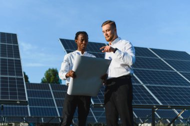 İki mühendis yenilenebilir enerji stratejisi planlıyor, güneşli bir günde güneş panelleri yakınındaki planları değerlendiriyorlar.