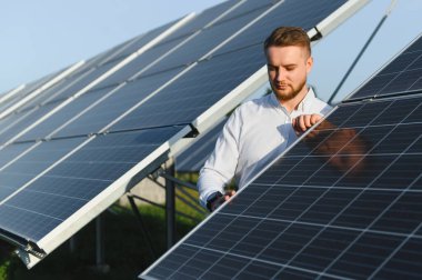 Beyaz gömlekli bir adam fotovoltaik çiftlikte güneş panellerini kontrol ediyor, sürdürülebilir temiz enerjiyi ve gelecekteki gücü simgeliyor.