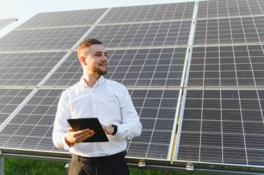 Elinde tablet olan beyaz tişörtlü, gülümseyen sakallı adam fotovoltaik bir fabrikada sürdürülebilir güneş enerjisi üretimini yönetiyor.