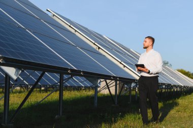 Genç adam, yenilenebilir enerji ve sürdürülebilirlik üzerine odaklanarak büyük bir güneş paneli dizisini incelerken dijital tablet kullanıyor.
