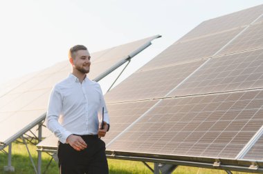 Gülümseyen mühendis elinde tablet, güneş panelleri arasında yürüyor, yenilenebilir enerji ve sürdürülebilir teknolojiyi temsil ediyor.